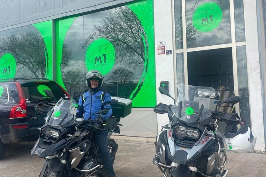 Une personne se tient à côté de deux motos devant un bâtiment doté de grandes fenêtres vertes et d’une enseigne sur laquelle on peut lire « M1 ». 4