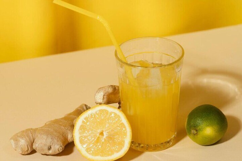 Et glas appelsinjuice med et sugerør, en halv citron, et stykke ingefær og en lime på et bord. 1
