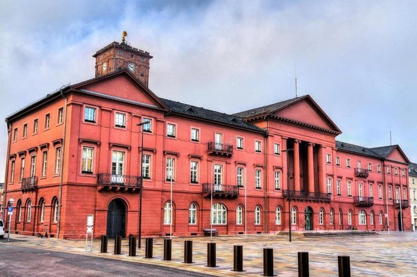 A red brick building with multiple windows and balconies, a clock tower, and a paved square in front. 3