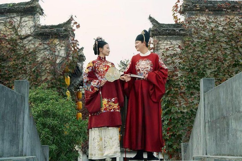 Two individuals in traditional attire standing in front of a traditional building with a wooden fence. 1