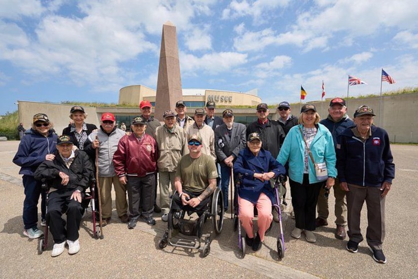Een groep mensen, waaronder mensen in rolstoelen, staat voor een monument met vlaggen op de achtergrond. 4