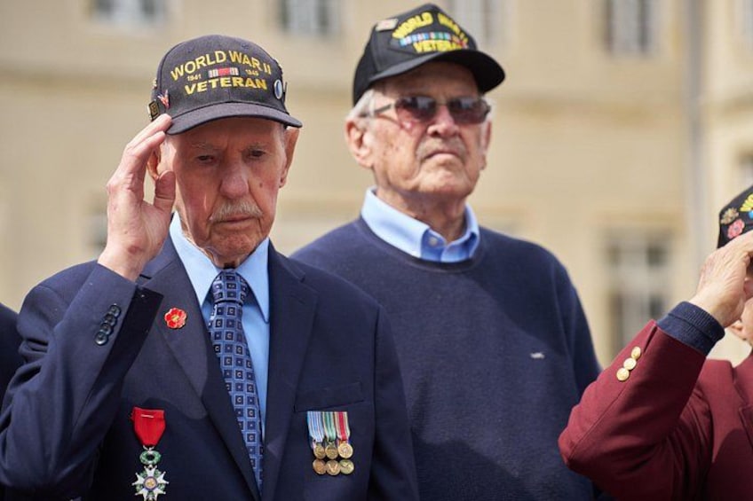 Twee mannen in militaire uniformen en petten, waarvan er één medailles draagt en de ander een pet met de tekst 'Wereldoorlog II Veteranen'. 3
