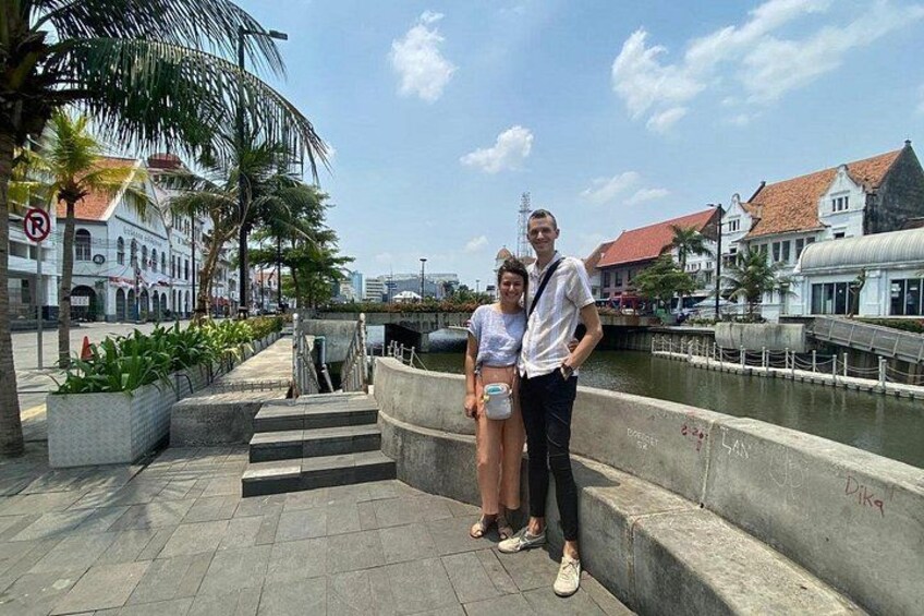 Ein Paar steht auf einer Uferpromenade, im Hintergrund sind Gebäude und Palmen zu sehen. 2