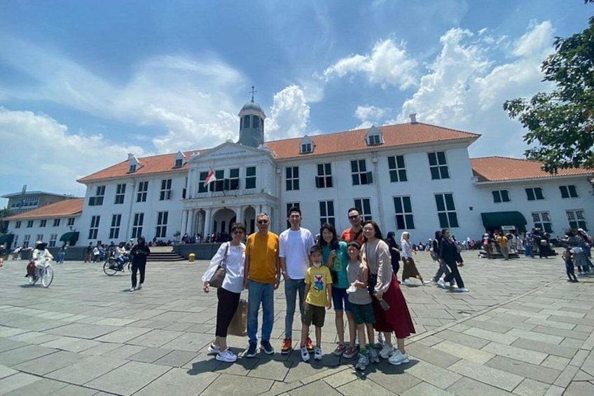 Eine Gruppe von Personen posiert vor einem großen, weißen Gebäude mit rotem Dach und einem Uhrturm. 1