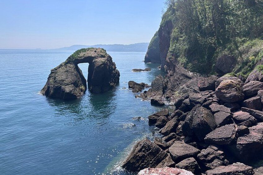 A coastal landscape with a prominent rock formation resembling an arch over the water, surrounded by rugged terrain and greenery. 3