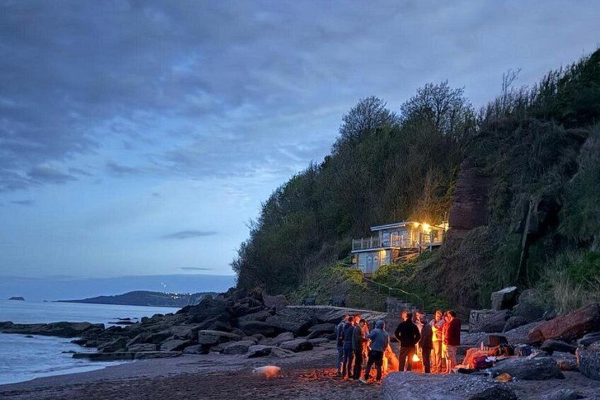 A coastal area with a house on a cliff, people gathered around a fire, and a rocky shoreline. 2