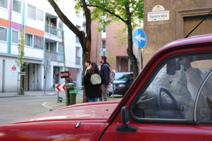 Un coche rojo estacionado en una calle con una señal direccional azul y un edificio que muestra el cartel 'Aurora utca'. 4