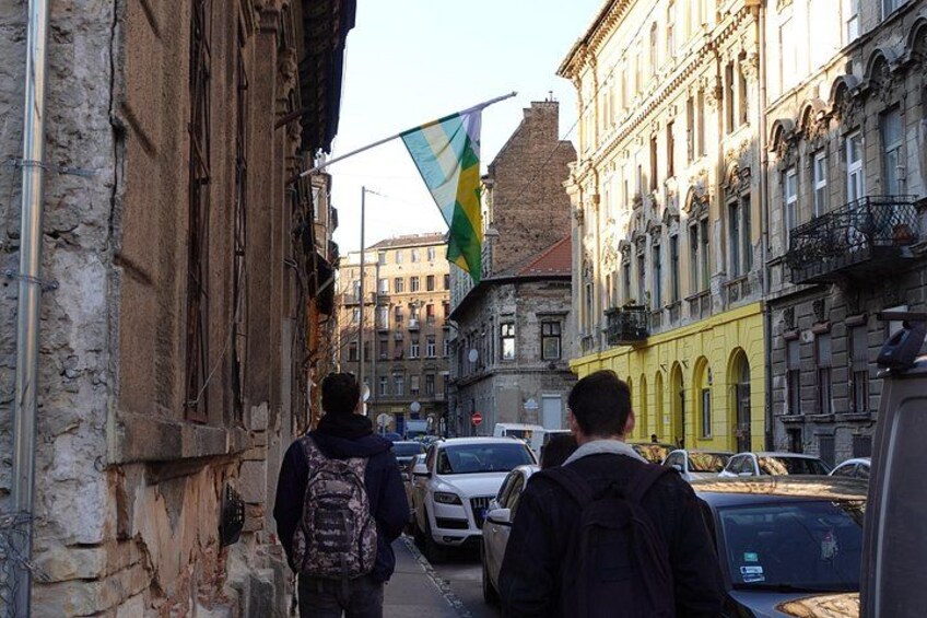 Una calle estrecha con edificios antiguos, coches aparcados y una bandera. 5