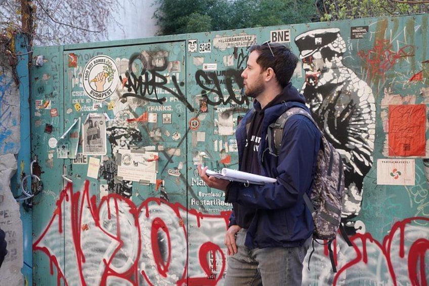 Un hombre de pie frente a un muro lleno de grafitis, con una mochila y un bloc de notas. 1