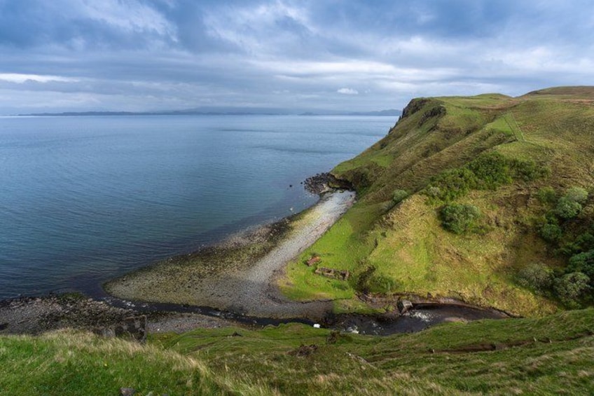 A coastal landscape with a grassy hill, a rocky shoreline, and a calm sea under a cloudy sky. 3