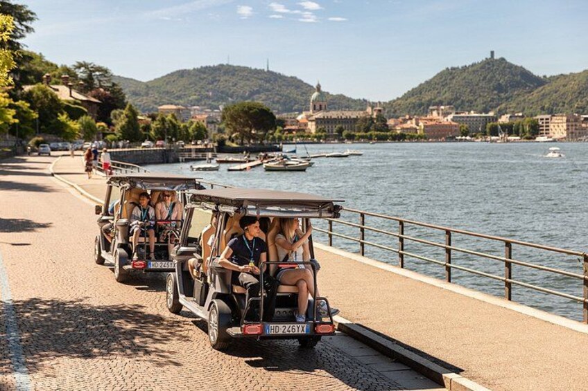 A group of people in a vehicle on a paved path by a body of water, with boats and hills in the background. 1
