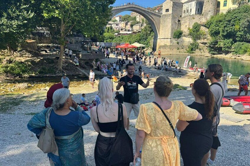 A group of people standing by a river with a bridge in the background. 1