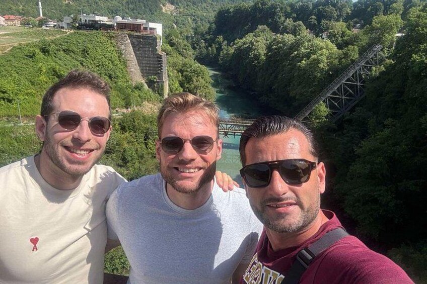 Three people standing with a bridge and greenery in the background. 2