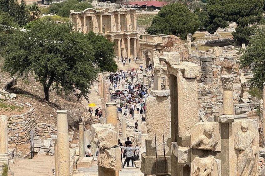 Ancient ruins with columns and statues, a crowd of visitors, and a large building in the background. 2