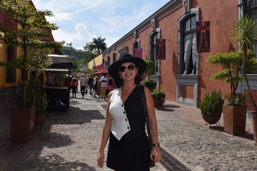 A cobblestone street with buildings, a person in a black dress and hat, and potted plants. 3