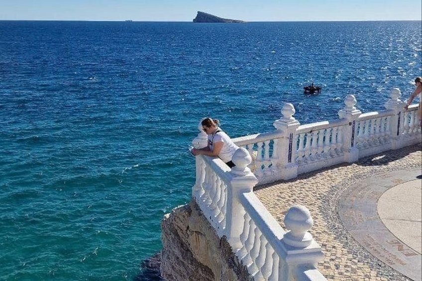 A coastal scene with a person standing on a white balustrade overlooking the sea, a rocky outcrop, and a distant island. 3