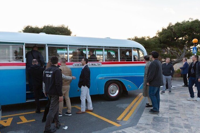 A blue bus with 'VINTA' and 'US MALTA' written on it, parked on a street with people boarding and alighting. 5