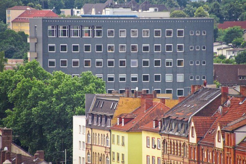 En moderne bygning med et gitter af vinduer, omgivet af farverige historiske bygninger med røde tegltage. 3