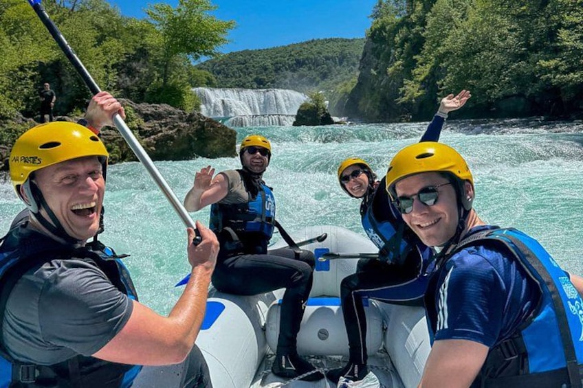 Eine Gruppe von Menschen treibt mit Rafts auf einem Fluss, im Hintergrund ein Wasserfall. 1