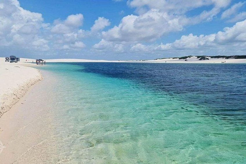 A beach with clear turquoise water, a sandy shore, and a few vehicles and people in the distance. 5