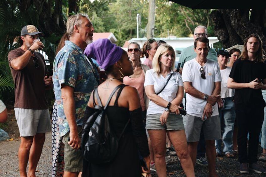 A group of people standing outdoors, some holding drinks, with trees and a building in the background. 2