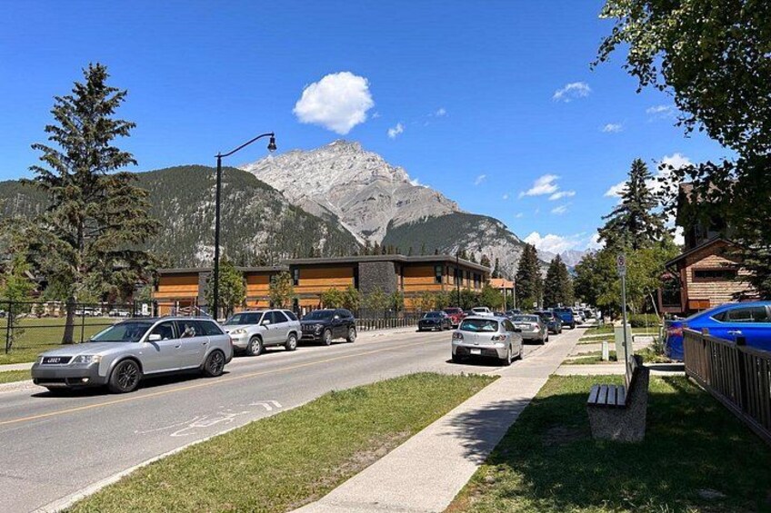 A road with parked cars, a building with a mountain backdrop, and a clear sky. 1