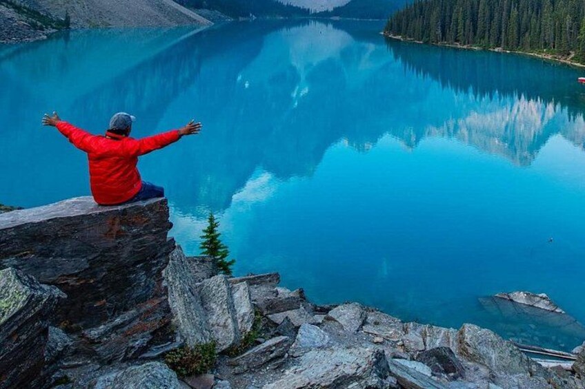 A person in a red jacket sitting on a rock with arms outstretched, overlooking a calm lake surrounded by rocky terrain and trees. 5
