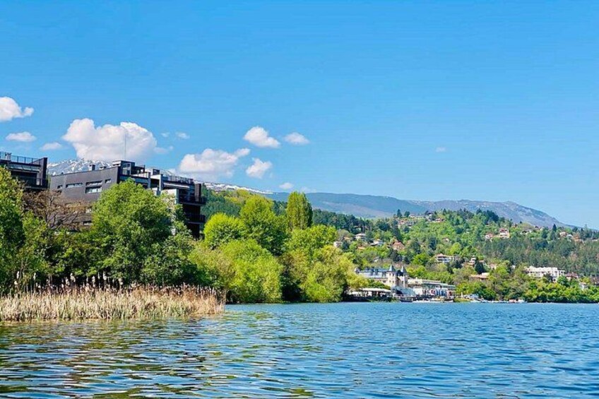 A lake with buildings on the left, trees in the foreground, and a hilly landscape in the background. 3