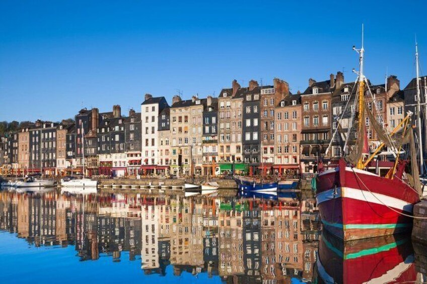 A harbor with a row of buildings reflecting on calm water, a red and white boat docked on the right, and several smaller boats in the water. 1