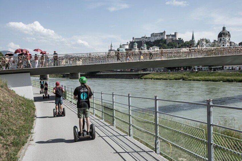 People riding Segways on a paved path beside a river, with a bridge and buildings in the background. 2