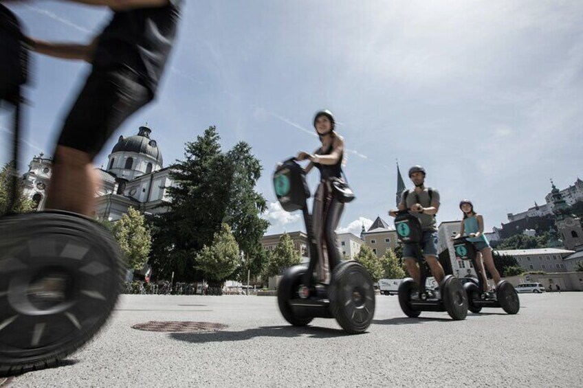 People riding Segways on a paved area with buildings and trees in the background. 3
