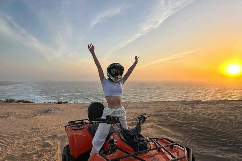 Eine Person auf einem Quad bei Sonnenuntergang mit erhobenen Armen, die auf den Ozean blickt. 1