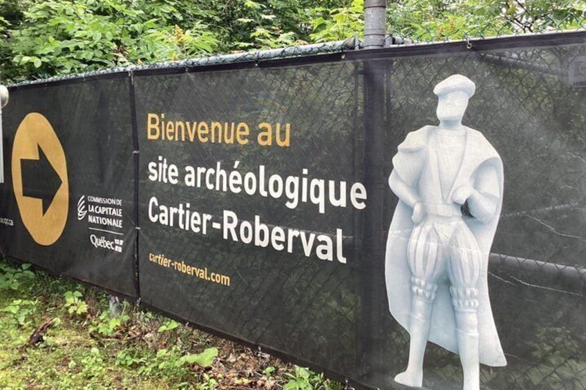 Señal de bienvenida al sitio arqueológico Cartier-Roberval, con una flecha indicadora y una estatua de una figura histórica. 1