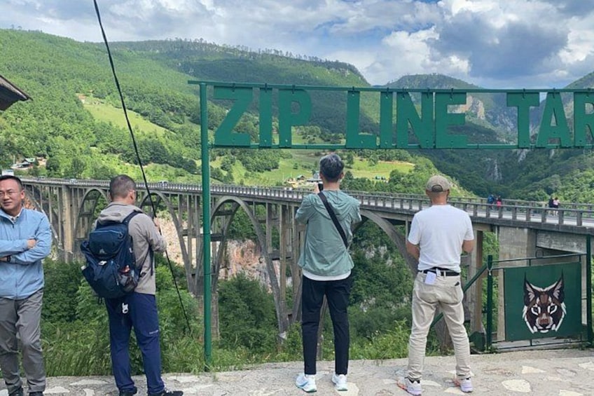 A group of people standing on a bridge with a green sign that reads 'ZIP LINE TAR.' 3