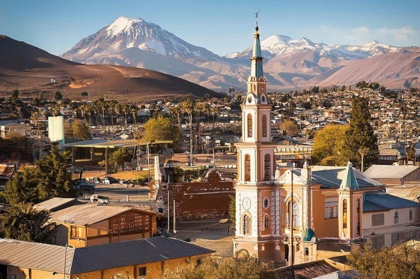 Un pueblo con una destacada torre de iglesia, una montaña nevada al fondo y una combinación de edificios residenciales y comerciales. 1