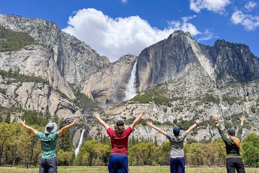 A group of people standing with arms raised in front of a waterfall and mountains. 1