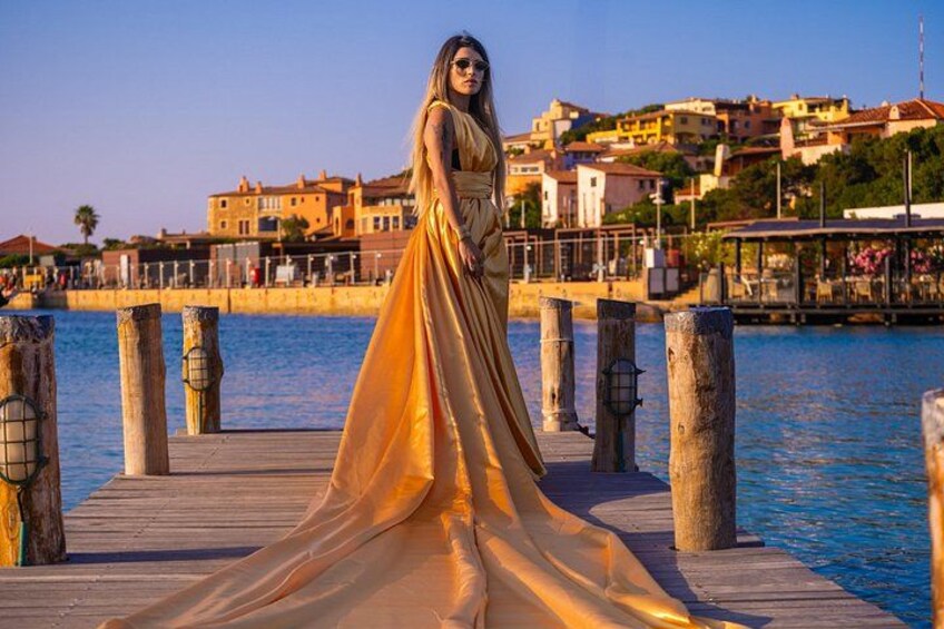 A woman in a flowing golden dress standing on a wooden pier with a coastal town in the background. 3