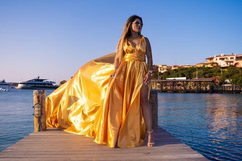 A woman in a golden dress standing on a wooden dock by a body of water with boats and buildings in the background. 5