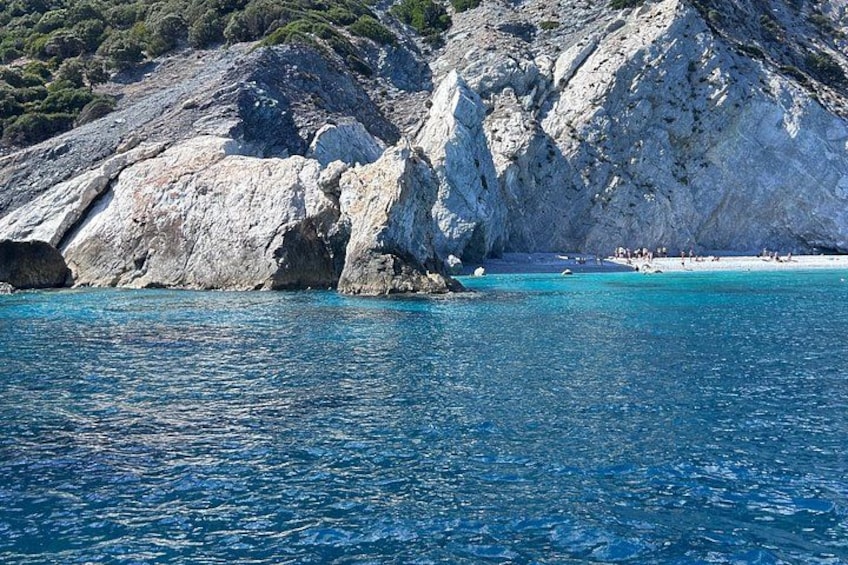 A coastal landscape with clear blue waters, rocky cliffs, and a beach with people. 2