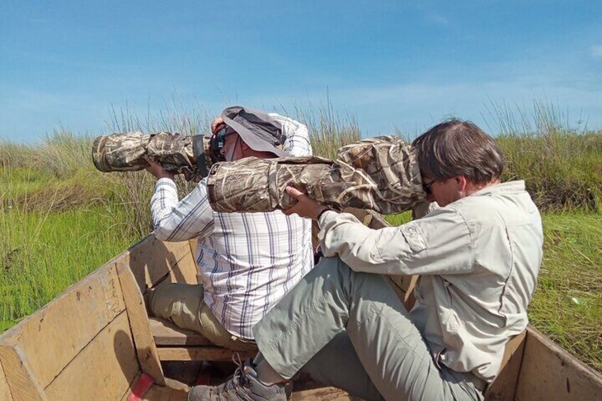 Two people in a wooden boat, one wearing camouflage gear and the other in a light shirt, with grass and sky in the background. 5