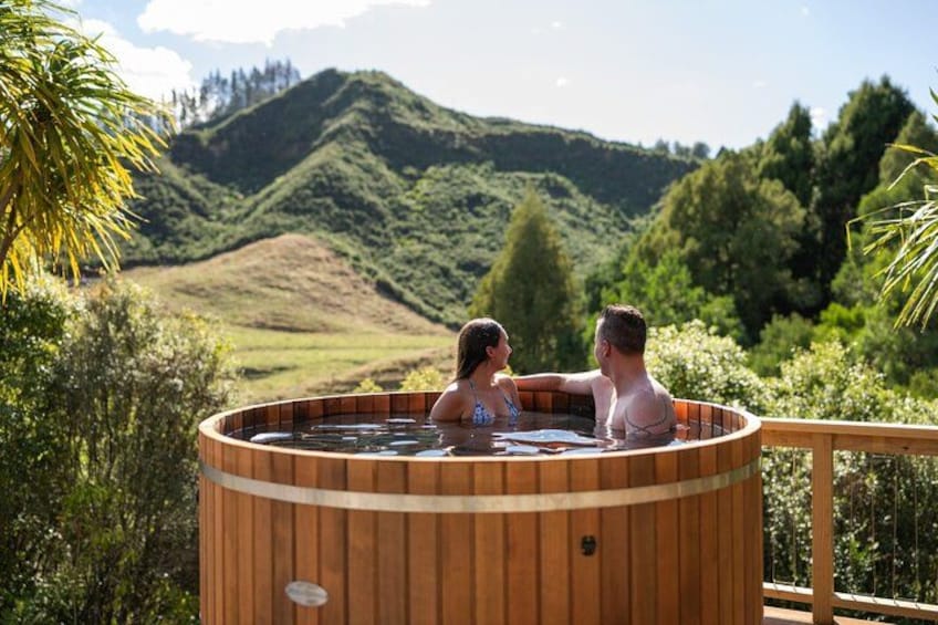 Un bain à remous en bois où se trouvent deux personnes, entouré d’une végétation luxuriante et avec des collines en arrière-plan. 2