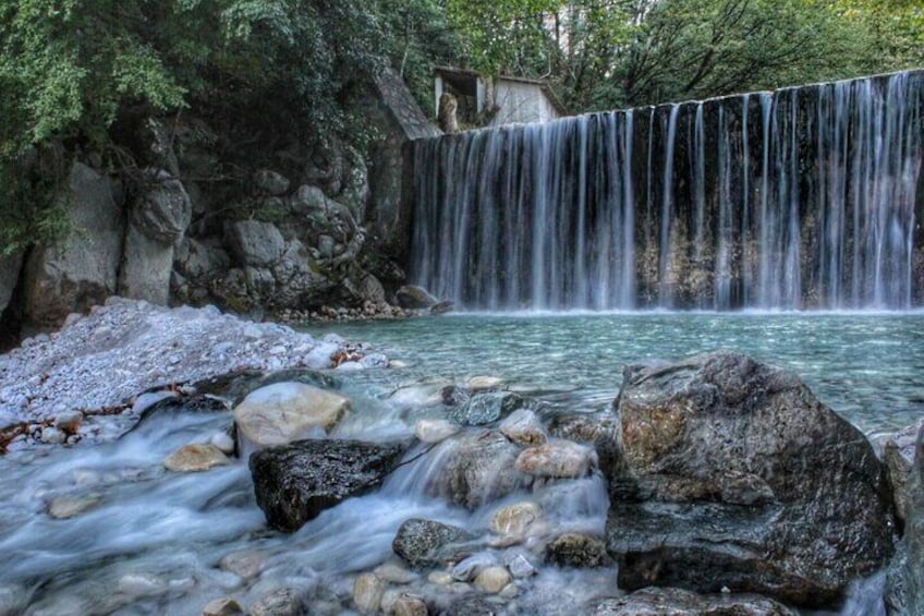 Een waterval stort over rotsen in een heldere beek, omgeven door bomen. 1