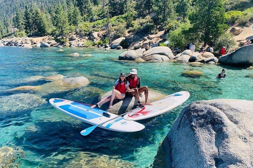 Two people sitting on surfboards in clear water with rocks and trees in the background. 1