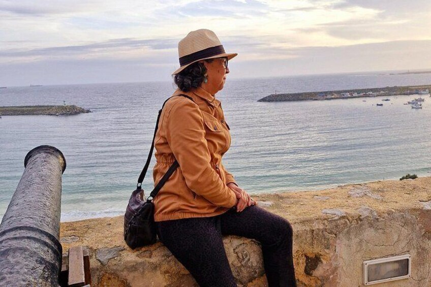 A person sitting on a stone wall overlooking a body of water with a lighthouse in the distance. 3
