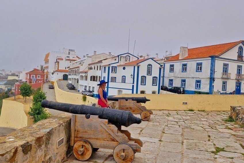 A cannon on a stone platform with a person in a red dress standing nearby. In the background, there are colorful buildings with blue and white facades. 5