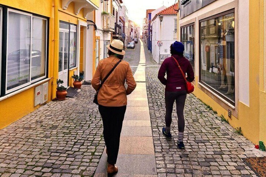 Two people walking down a cobblestone street lined with buildings. 2