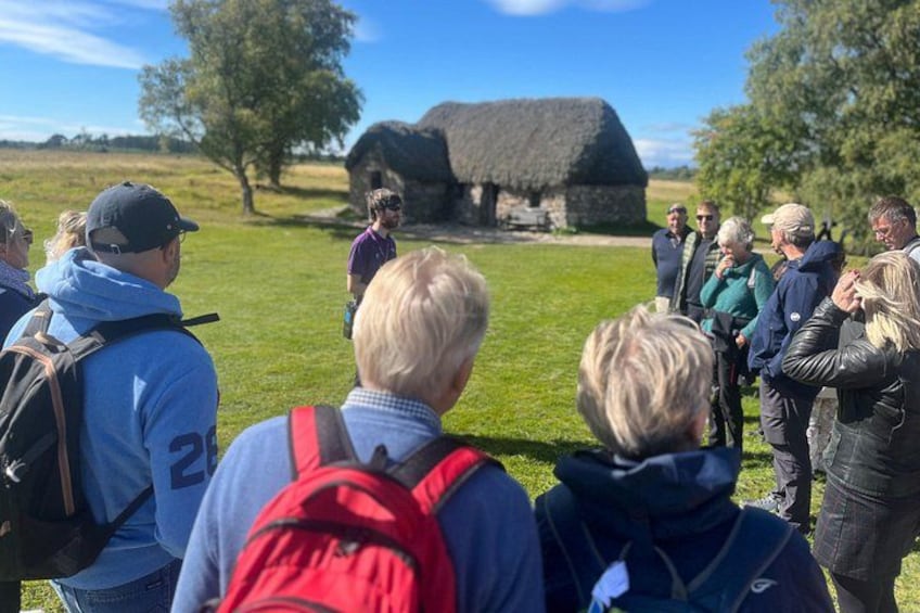 A group of people standing outdoors, a thatched-roof building, and a grassy field. 4