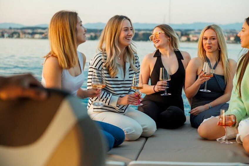 Een groep mensen zit op een boot, drinken in de hand, met een wateroppervlak en een verre kustlijn op de achtergrond. 5