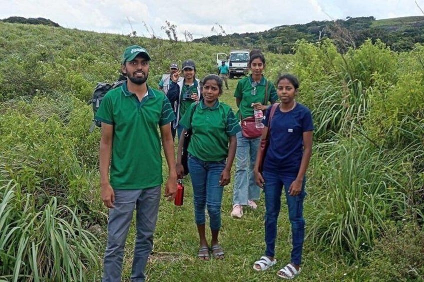 A group of people in green shirts standing in a grassy area with a vehicle in the background. 3