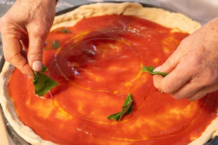 Manos extendidas untando salsa de tomate sobre la masa de una pizza con hojas de albahaca. 5